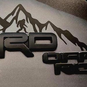 May include: Black TRD Off Road emblem with a mountain range silhouette and a Bigfoot figure. The emblem is a matte black finish, perfect for off-road enthusiasts.