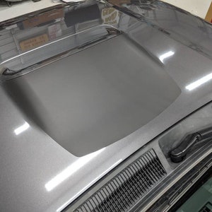 May include: A gray car hood with a black vinyl hood scoop. The hood scoop is a popular modification for cars, as it can improve airflow and reduce drag.