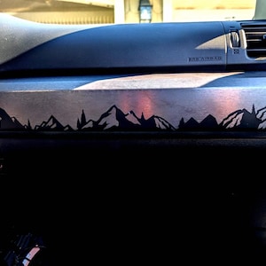 May include: A black and silver car dashboard panel with a mountain range silhouette design. The design features a series of black mountain peaks and trees against a silver background. The dashboard also has an "SRS AIRBAG" label.