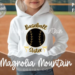 May include: White hoodie with a black and gold baseball graphic that says "Baseball Sister". The hoodie has a front pocket and a hood. The text "Magnolia Mountain Design Co" is at the bottom.