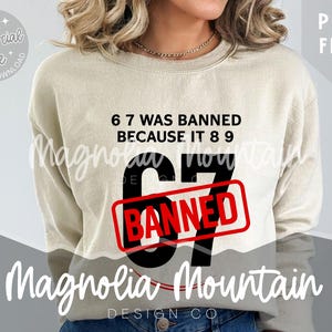 May include: A beige sweatshirt with the text "6 7 WAS BANNED BECAUSE IT 8 9". The number "67" is in large black letters with a red "BANNED" stamp over it. The words "Magnolia Mountain DESIGN CO" are also visible.