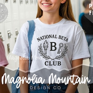 May include: A white t-shirt with the words "NATIONAL BETA CLUB" in black. The design features a torch, the letter "B", and a key, surrounded by a laurel wreath. The shirt is worn by a person with a blue shoulder bag.