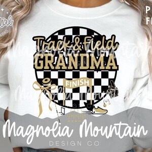 May include: White sweatshirt with a graphic that says "Track & Field Grandma." The design features a black and white checkered circle with gold accents, a finish line banner, and track and field-related icons. The text "Magnolia Mountain Design Co" is at the bottom.