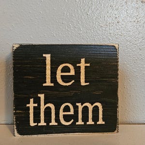 May include: A black wooden sign with white lettering that reads "let them".