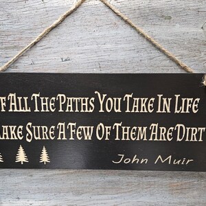 May include: Black wooden sign with white text that reads "Of all the paths you take in life make sure a few of them are dirt" and "John Muir". The sign has three small pine tree silhouettes.
