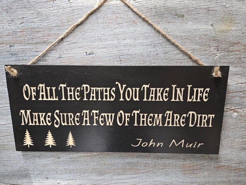 May include: Black wooden sign with white text that reads "Of all the paths you take in life make sure a few of them are dirt" and "John Muir". The sign has three small pine tree silhouettes.