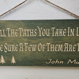May include: A green wooden sign with a white inscription that reads "OF ALL THE PATHS YOU TAKE IN LIFE MAKE SURE A FEW OF THEM ARE DIRT" and the name "John Muir" at the bottom. Three small pine trees are depicted below the text.