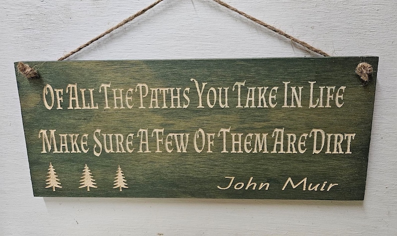May include: A green wooden sign with a white inscription that reads "OF ALL THE PATHS YOU TAKE IN LIFE MAKE SURE A FEW OF THEM ARE DIRT" and the name "John Muir" at the bottom. Three small pine trees are depicted below the text.