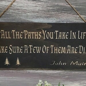 May include: A dark brown wooden sign with the quote "Of all the paths you take in life make sure a few of them are dirt" by John Muir. The sign has three small pine tree silhouettes.