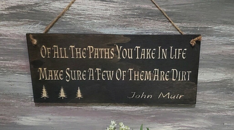 May include: A dark brown wooden sign with the quote "Of all the paths you take in life make sure a few of them are dirt" by John Muir. The sign has three small pine tree silhouettes.