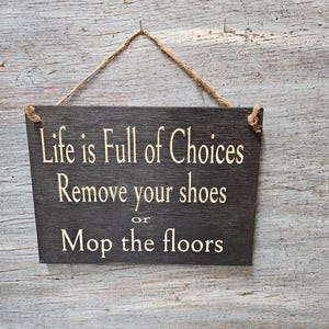 May include: A dark brown wooden sign with white text that reads "Life is Full of Choices Remove your shoes or Mop the floors". The sign is hanging from a piece of twine.