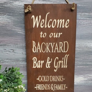 Puede incluir: Se&ntilde;al de madera marr&oacute;n r&uacute;stica con texto blanco que dice "Bienvenido a nuestro Bar y Grill del patio trasero - BEBIDAS FR&Iacute;AS - AMIGOS Y FAMILIA - EXCELENTE COMIDA".