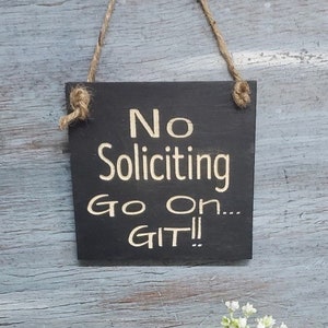 May include: A black wooden sign with white text that reads "No Soliciting Go On... GIT!" The sign is hanging from a piece of twine.
