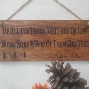 May include: Wooden sign with the quote "Of all the paths you take in life make sure a few of them are dirt" attributed to John Muir. The sign has a rustic brown finish and three small pine tree silhouettes.