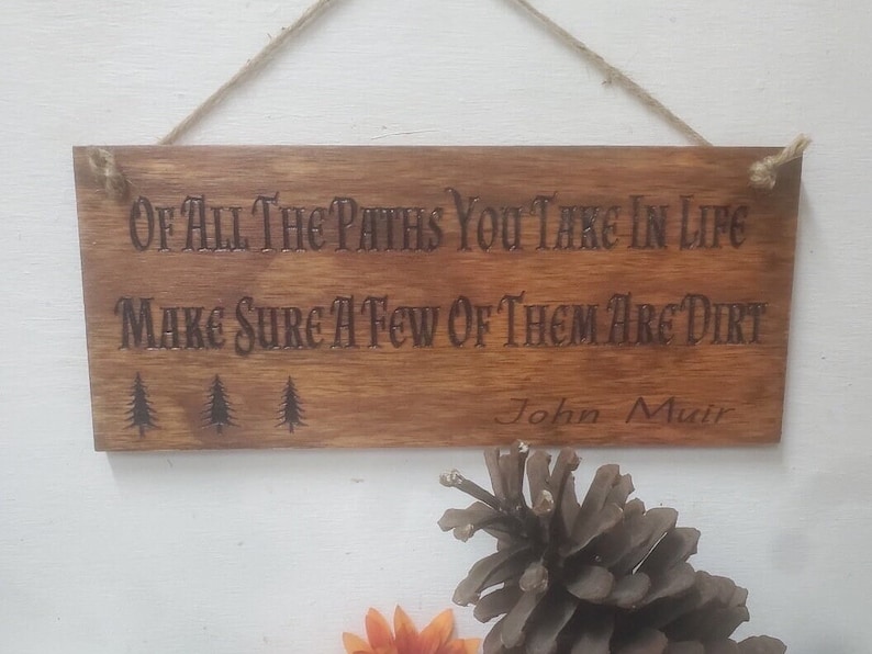 May include: Wooden sign with the quote "Of all the paths you take in life make sure a few of them are dirt" attributed to John Muir. The sign has a rustic brown finish and three small pine tree silhouettes.