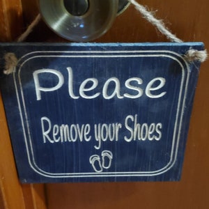 May include: A blue wooden sign with white text that reads "Please Remove your Shoes" with a pair of flip-flop icons below the text. The sign is hanging on a wooden door with a brass doorknob.