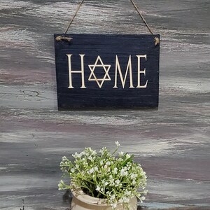 May include: A black wooden sign with the word "HOME" in white lettering and a gold Star of David in the center. The sign is hanging from a string and is in front of a gray wooden background.