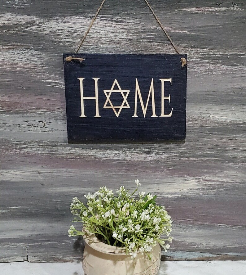 May include: A black wooden sign with the word "HOME" in white lettering and a gold Star of David in the center. The sign is hanging from a string and is in front of a gray wooden background.