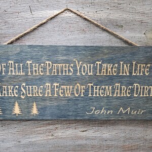 May include: A rustic wooden sign with a blue-grey stain. The sign has a quote from John Muir: "Of all the paths you take in life make sure a few of them are dirt."  The quote is in a light brown color and there are three stylized pine trees to the left of the quote.