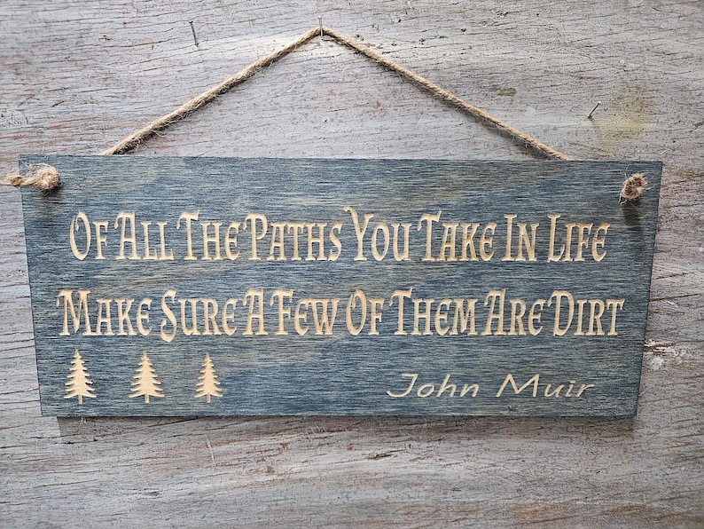 May include: A rustic wooden sign with a blue-grey stain. The sign has a quote from John Muir: "Of all the paths you take in life make sure a few of them are dirt."  The quote is in a light brown color and there are three stylized pine trees to the left of the quote.