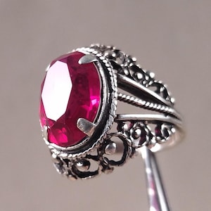 Może przedstawiać: Srebrny pierścionek z owalnym, głęboko czerwonym kamieniem szlachetnym. Pierścionek ma ozdobny, filigranowy wzór wokół kamienia i na obrączce. Kamień szlachetny jest centralnym punktem, osadzonym w szczegółowej, stylizowanej oprawie.