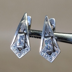 Puede incluir: Un par de pendientes plateados con un diseño geométrico. Cada pendiente presenta una piedra rectangular central y está acentuado con piedras más pequeñas. Los pendientes están sujetos por una herramienta plateada sobre un fondo neutro.