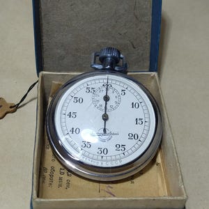 May include: A vintage stopwatch with a white face, black numerals, and a silver-coloured metal casing. It is displayed open inside a blue box with a beige interior. A small tag is attached.