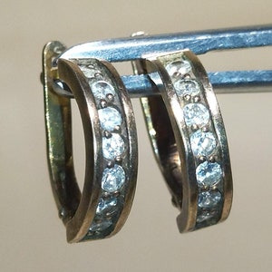 以下が含まれることがあります： 小さな透明な宝石がちりばめられた、金色のフープピアス。ニュートラルな背景に対して金属製のツールで保持されており、ジュエリーのデザインと職人技が際立っています。