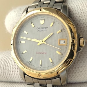 May include: An Orient Automatic wristwatch with a silver-toned dial, gold-toned bezel, and a titanium link bracelet. The dial features gold-toned hour markers, gold-toned hands, a date window at the three o'clock position, and the text TITANIUM.