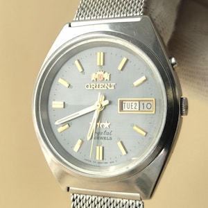 May include: A silver-tone Orient wristwatch with a light blue dial, gold-tone hour markers, and hands. The watch features a day-date window at the three o'clock position displaying TUE 2 10 and a stainless steel mesh strap with a metallic finish.
