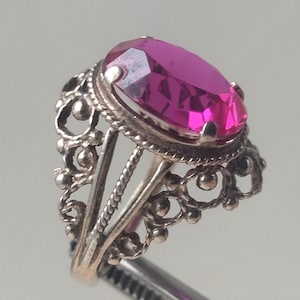 May include: A vintage-style ring with a large, oval-cut, magenta gemstone. The ring features an ornate, filigree design in a silver-tone metal. The gemstone is held in place by a bezel setting, and the ring is held by a pair of tweezers.