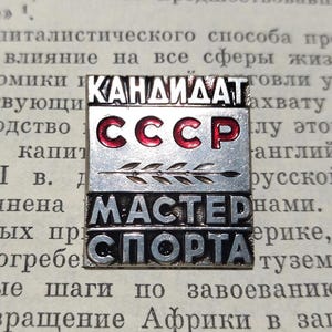 Könnte beinhalten: Ein silberfarbener Anstecker mit den kyrillischen Wörtern "КАНДИДАТ СССР" in Rot und "МАСТЕР СПОРТА" darunter. Der Anstecker hat ein Zweig-Design und ist vor einem Hintergrund aus gealtertem Papier platziert.