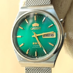May include: An Orient wristwatch with a vibrant green dial, gold-tone hour markers, and gold hands. The watch features a day-date window displaying THU 6, a three-star logo, and the text Crystal 21 Jewels. It has a silver-tone metal mesh strap.
