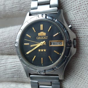 Op de afbeelding: Een zilverkleurig Orient horloge met een zwarte wijzerplaat en gouden accenten. Het horloge heeft een datumweergave en een zilveren band. Het horloge wordt getoond op een gehandschoende hand.