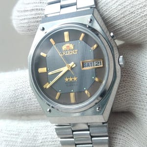 May include: A silver-toned Orient watch with a gray face and gold accents. The watch displays the day and date, with the day reading "SAT 21". The watch has a silver band and is held by a gloved hand.