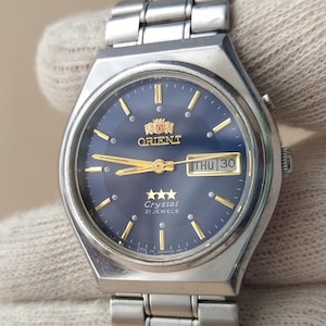 May include: A silver-tone stainless steel Orient Crystal watch with a dark blue circular dial, gold-tone hands and hour markers, and a day-date window at the three o'clock position. The watch features a metal link bracelet and a three-star logo.