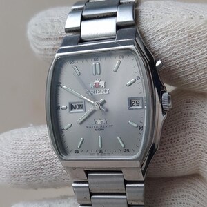 May include: A silver-toned Orient wristwatch with a rectangular case and a matching stainless steel link bracelet. The dial features a light silver finish, silver hour markers, a day-date window at the three o'clock position, and the Orient logo.