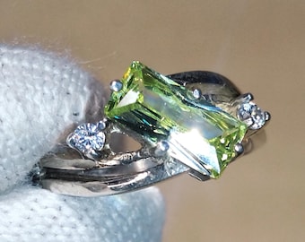 May include: A silver ring featuring a rectangular, light green gemstone as the primary focal point. The ring is accented with smaller, clear gemstones. The design is elegant and modern, suitable for various occasions.