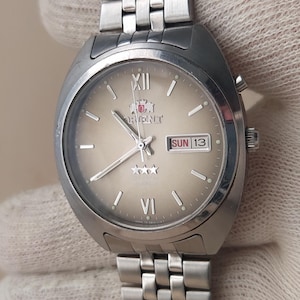 May include: A silver-toned analogue wristwatch with a metallic link bracelet. The watch features a champagne-coloured dial with silver hour markers, Roman numerals at twelve and six, a day-date window at three, and three small stars at six.