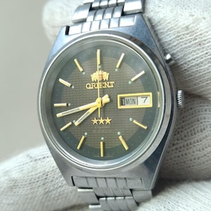 May include: Silver-toned Orient watch with a dark green face and gold accents. The watch features a day and date display, along with gold hands and hour markers. The watch has a silver band and is a classic timepiece.