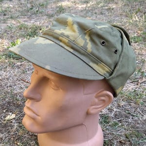 Puede incluir: Gorra de camuflaje verde oliva con visera y orejeras. El sombrero presenta patrones beige y marrón. El sombrero se muestra en una cabeza de maniquí.