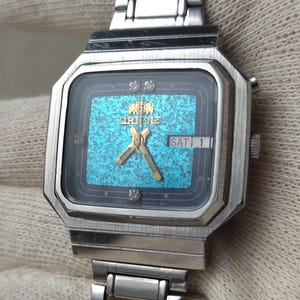 May include: A vintage Orient watch with a silver-tone rectangular case and a matching link bracelet. The watch face features a turquoise textured background, gold hands, and a day/date display. The watch is shown against a neutral background.
