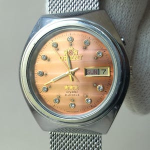 Op de afbeelding: Een vintage Orient horloge met een koperkleurige wijzerplaat, kristallen accenten en gouden wijzers. Het horloge toont "SUN 7" en "Crystal 21 Jewels". Het heeft een zilveren mesh band.