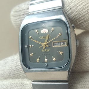 May include: A silver-toned Orient wristwatch featuring a square-shaped case and a matching metal link bracelet. The dark gray dial displays gold-colored hour markers, gold hands, a day-date window at the three o'clock position, and the Orient logo.