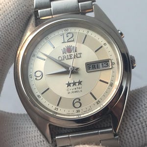 May include: Silver-toned Orient watch with a cream-colored face, featuring the day and date. The watch has a stainless steel band and a classic design. The face displays the Orient logo, the day of the week, and the time.