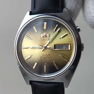 May include: A silver-toned Orient AAA Crystal automatic wristwatch with a black leather band. The watch face is gold with black hands and markers. The day and date are displayed at the 3 o'clock position.