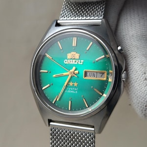 May include: A silver-toned Orient watch with a green dial and a mesh band. The watch face displays the Orient logo, gold-colored hands, and the day and date. The watch is held by a gloved hand.