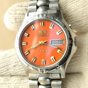 May include: An Orient wristwatch with a bright orange dial, gold-tone hands, and hour markers. The watch features a stainless steel case and link bracelet, a day-date window at the three o'clock position, and three small stars below the logo.