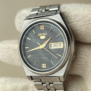 May include: A silver-toned Seiko wristwatch with a dark gray face and gold hands. The watch features a day and date display, with the day reading "LUN 4". The watch has a stainless steel band and a textured dial.
