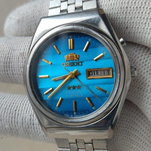 May include: A silver-toned Orient watch with a blue dial and a stainless steel band. The watch face features gold-colored hands and markers, and a day-date display. The watch is a classic design.